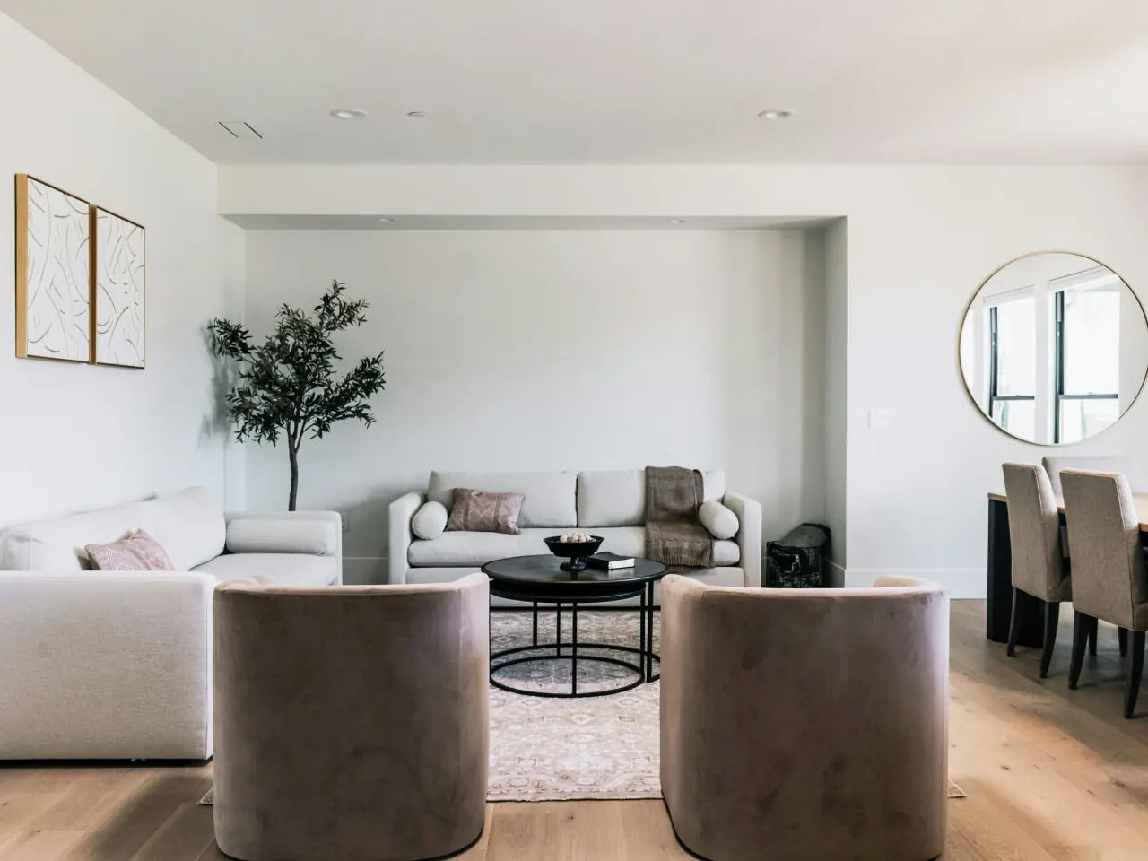 Model home living room with pale oak floors, two blush velvet swivel chairs facing a cream sofa, a round black coffee table and a potted olive tree