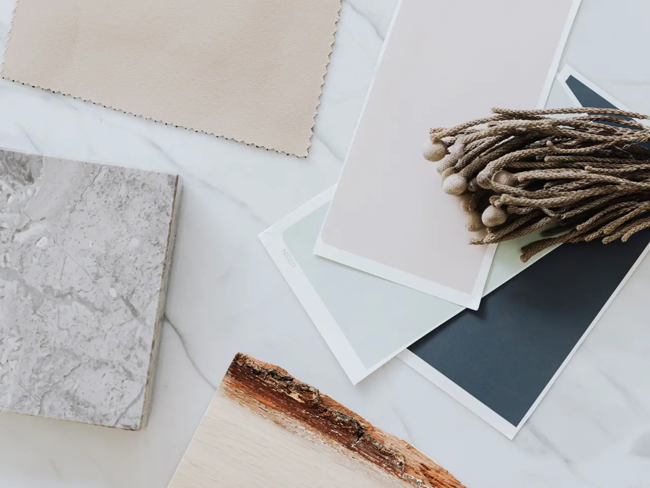 Finish samples laid out on white marble: a grey stone tile, paint chips in blush, sage and slate, a beige fabric swatch and a raw-edge oak board