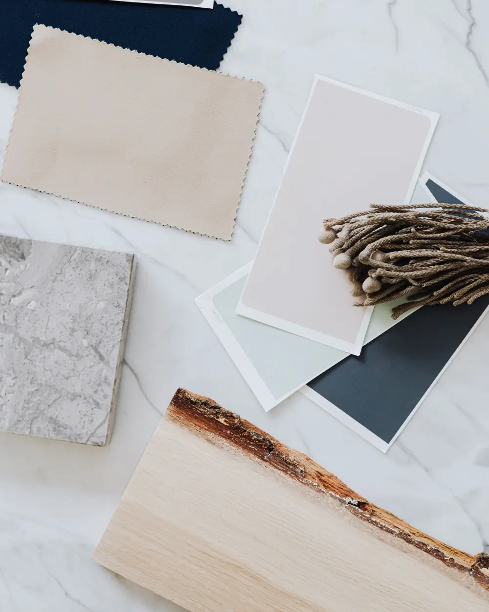 Finish samples laid out on white marble: a grey stone tile, paint chips in blush, sage and slate, a beige fabric swatch and a raw-edge oak board
