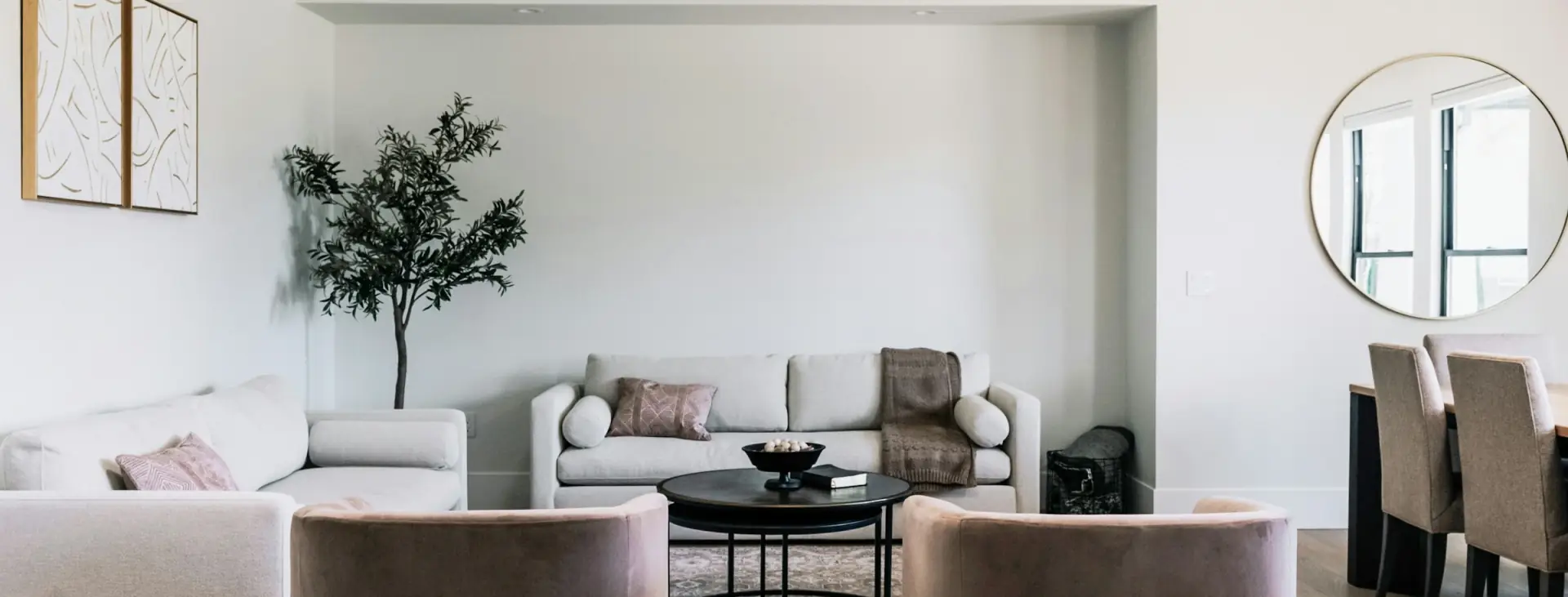 Model home living room with pale oak floors, two blush velvet swivel chairs facing a cream sofa, a round black coffee table and a potted olive tree