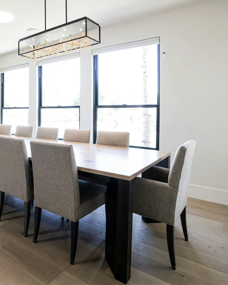 Corner of a bright dining room with black-framed windows, a long oak table, grey upholstered chairs and a glass box chandelier