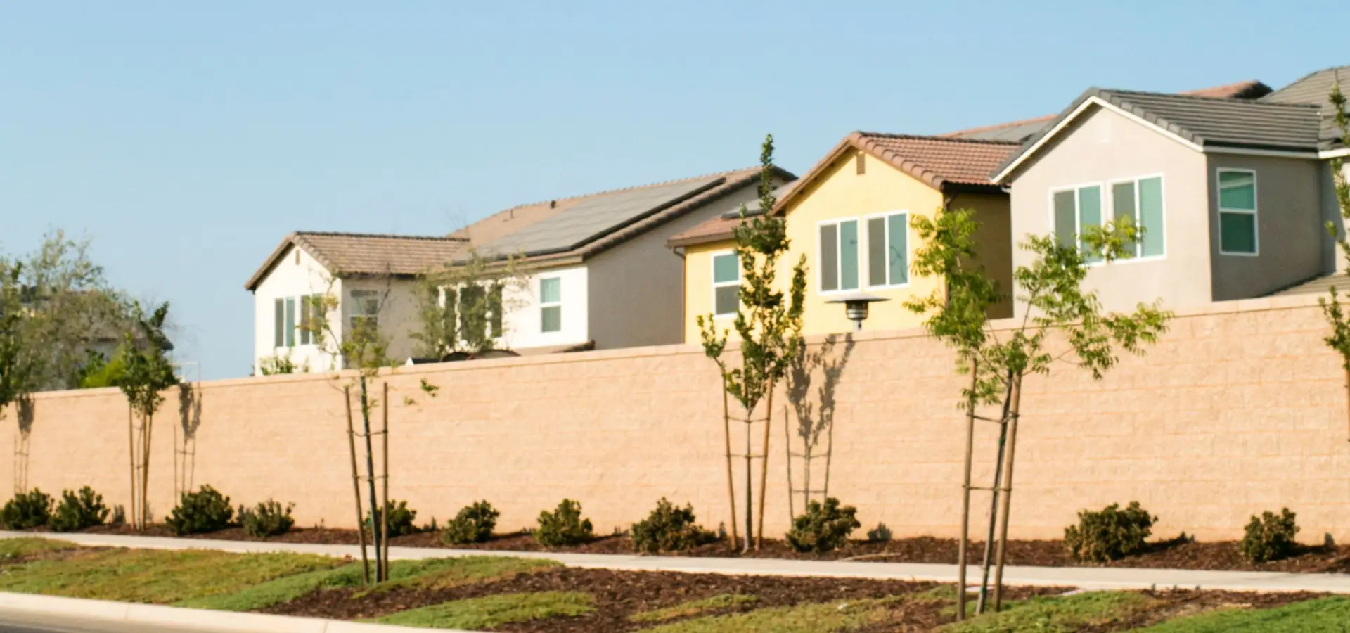 A new stucco perimeter wall lined with staked young trees and fresh mulch beds along a sidewalk, with two-storey tile-roof homes rising behind it