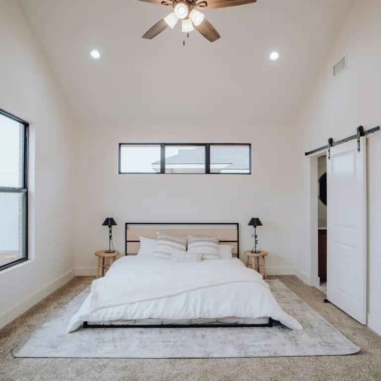 Primary bedroom under a vaulted ceiling with a ceiling fan, a clerestory window, a white-made bed on a grey rug and a sliding barn door