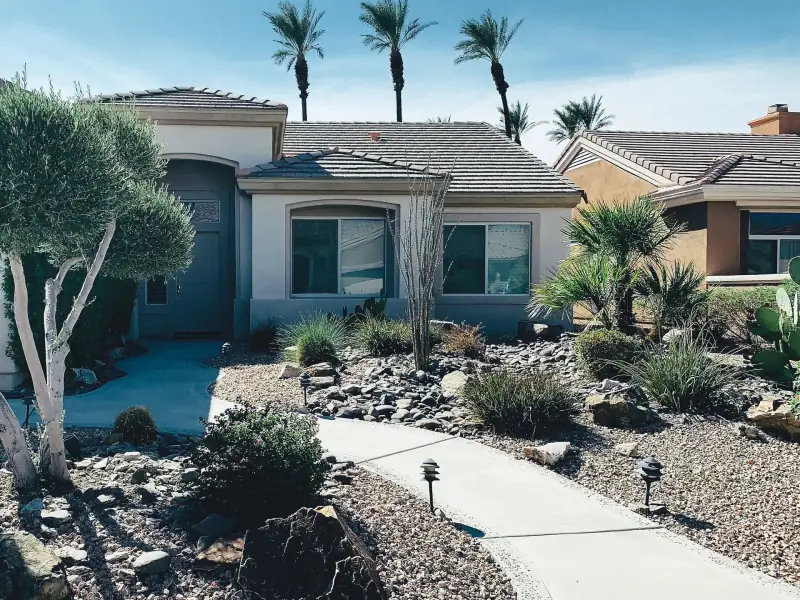 Single-storey stucco home with a tile roof, a curving concrete path and desert landscaping of gravel, boulders, agave and a pale-barked olive tree