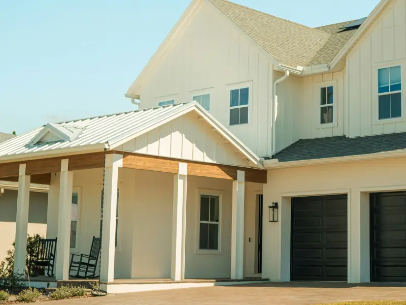 White board-and-batten farmhouse front with a metal-roofed porch, a cedar beam, black rocking chairs and dark garage doors