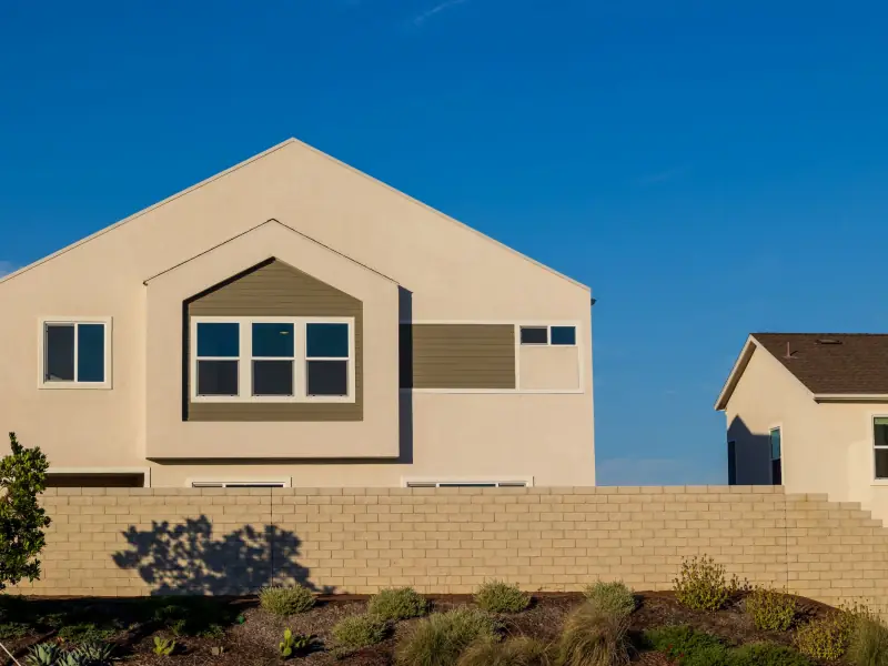 Modern two-storey stucco home with a gabled front and a framed window bay, seen above a block garden wall planted with drought-tolerant shrubs