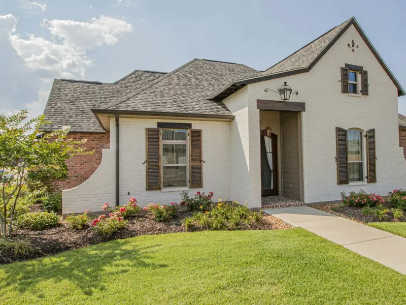 Painted white brick home with dark shutters, a lantern over the recessed entry, red brick side wings and rose beds along the walk