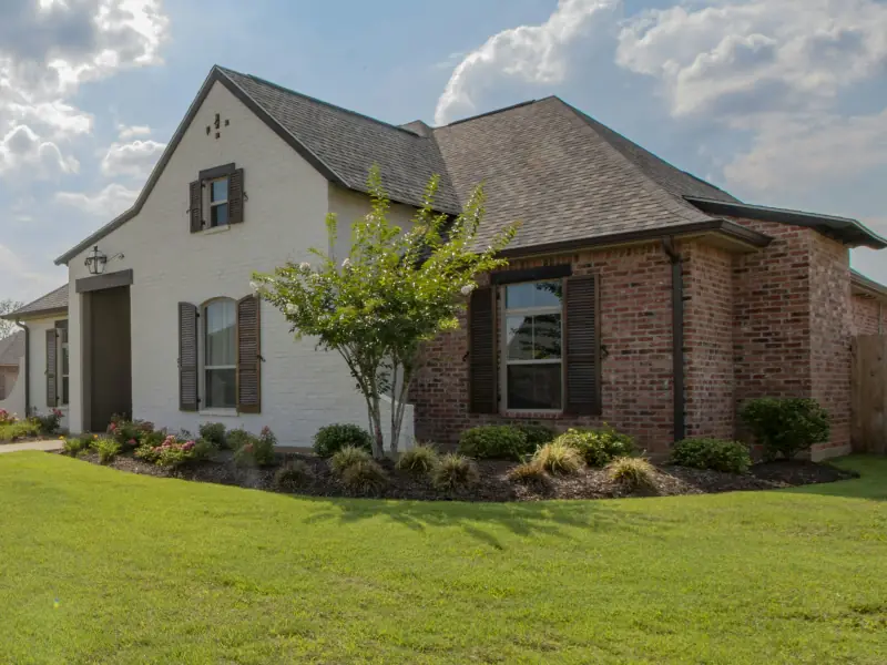 Corner lot home in white-painted and red brick with dark shutters, a young crape myrtle and a wide fresh lawn