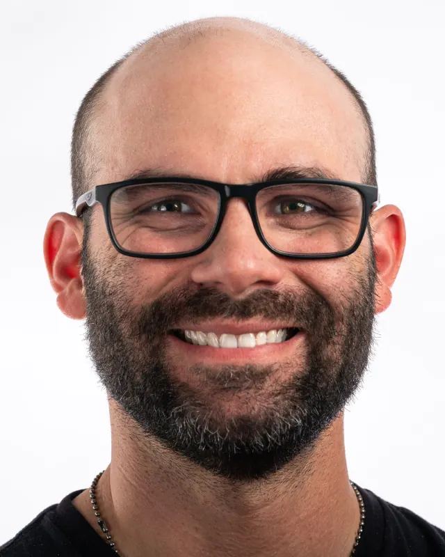Bald man with a full beard and black-framed glasses, smiling broadly, against a white backdrop