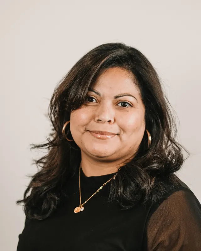 Woman with long dark wavy hair and gold hoop earrings in a black top, smiling against a light grey wall